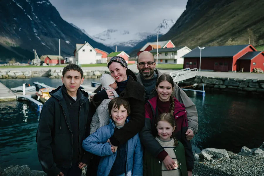 Szene aus „Fjord“: Eine Familie mit fünf Kindern steht lächelnd an einem kleinen Hafen, dahinter rote und weiße Holzhäuser und schneebedeckte Berge